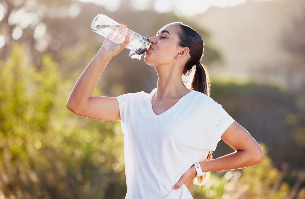 A woman takes a drink of water from a bottle while standing outdoors, with one hand on her hip, after exercising - Chiropractor Meridian Idaho A woman takes a drink of water from a bottle while standing outdoors, with one hand on her hip, after exercising - Chiropractor Meridian Idaho