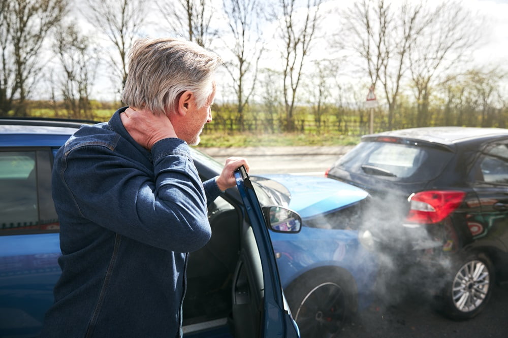 A man holds his neck in pain as he exits his car following a rear-end collision, with two damaged cars visible on the road - Chiropractor Meridian Idaho A man holds his neck in pain as he exits his car following a rear-end collision, with two damaged cars visible on the road - Chiropractor Meridian Idaho